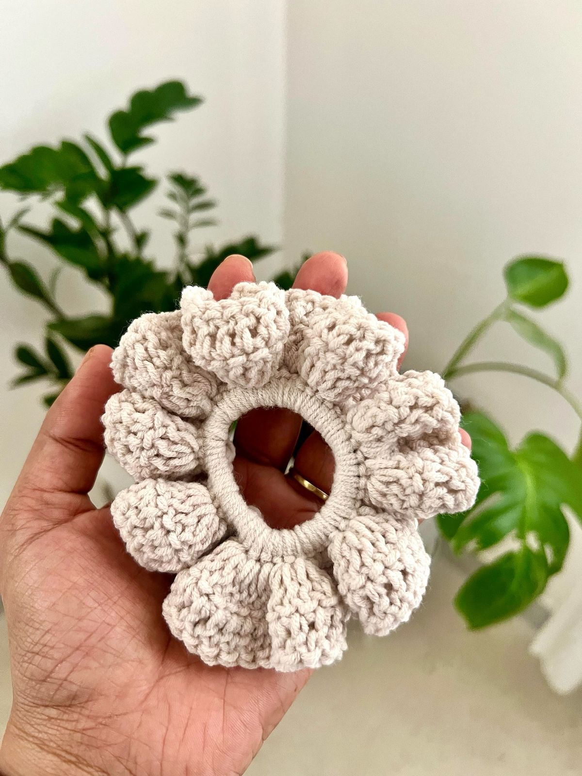 A hand holding a finished cream crochet scrunchie, with green houseplants in the background.