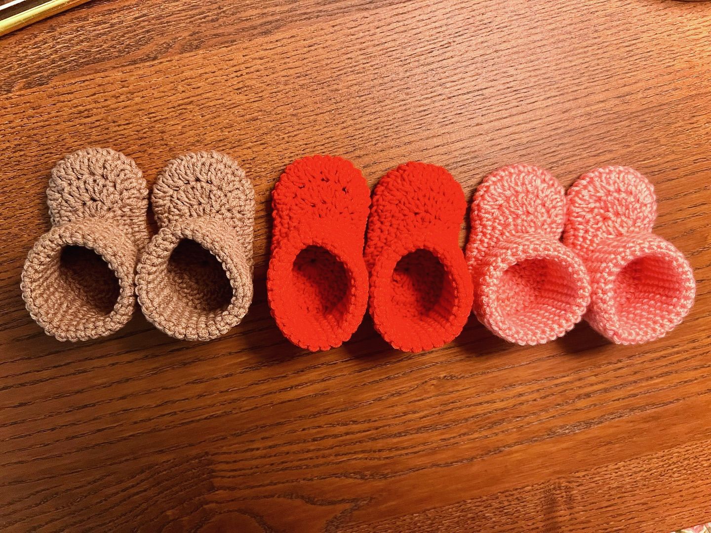 Three pairs of hand-crocheted baby booties in tan, red and pink, laid out on a wooden table.