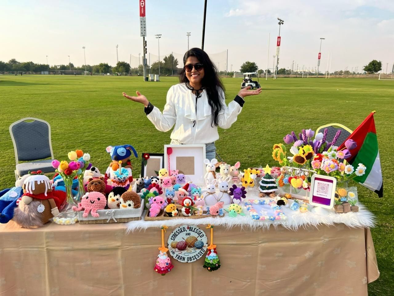 Shilpa smiling at her Loomé market stall on a grass field, with the UAE flag and colorful crochet flowers on the table.